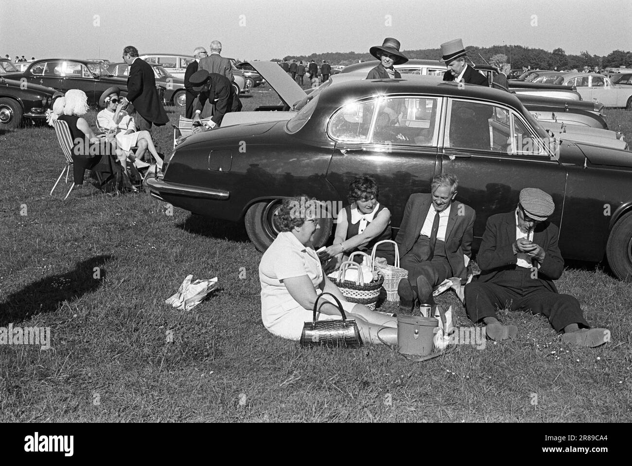 Oberschicht der Arbeiterklasse, Hut und Schwanz, Stoffkappe und Pfeife, Derby Day. Londoner verbringen einen Tag bei den Rennen, genießen ein Picknick im Freien auf dem Parkplatz, geschützt vor der Brise und warm in der Sonne, lehnen sie sich gegen einen zurückkehrenden Rennfahrer Jaguar. Epsom Downs, Surrey, England, 3. Juni 1970. HOMER SYKES AUS DEN 1970ER JAHREN Stockfoto