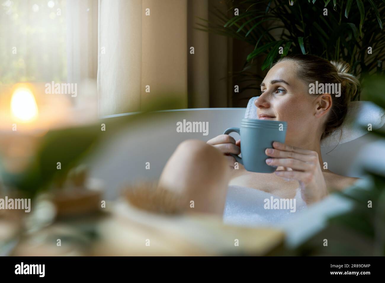 Frau, die sich in der Badewanne entspannt und einen Kaffee im Badezimmer zu Hause trinkt. Aus dem Fenster sehen Stockfoto