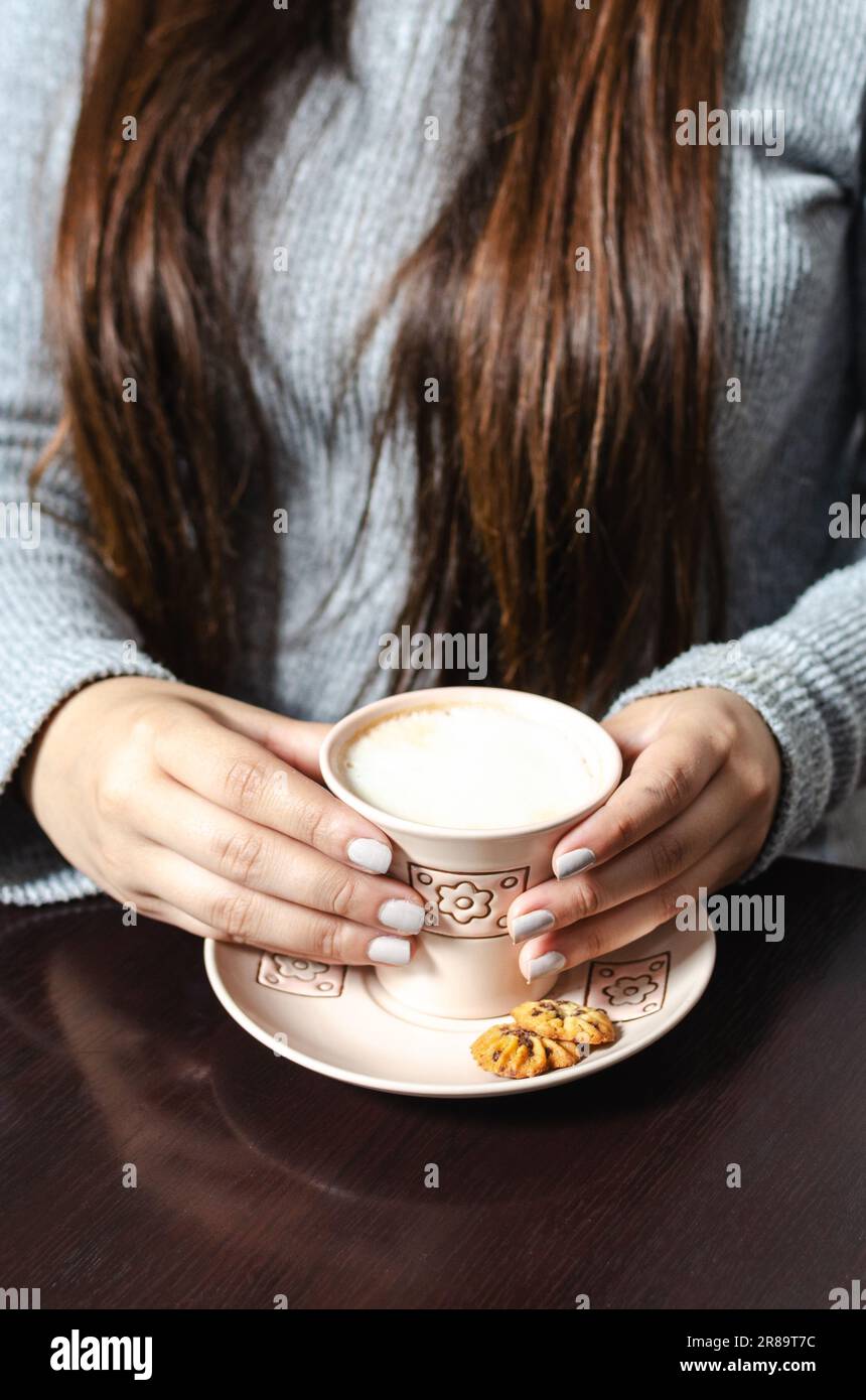 Frauenhände mit einer Tasse Kaffee mit Milch, dazu Mini-Kekse auf einem Holztisch Stockfoto