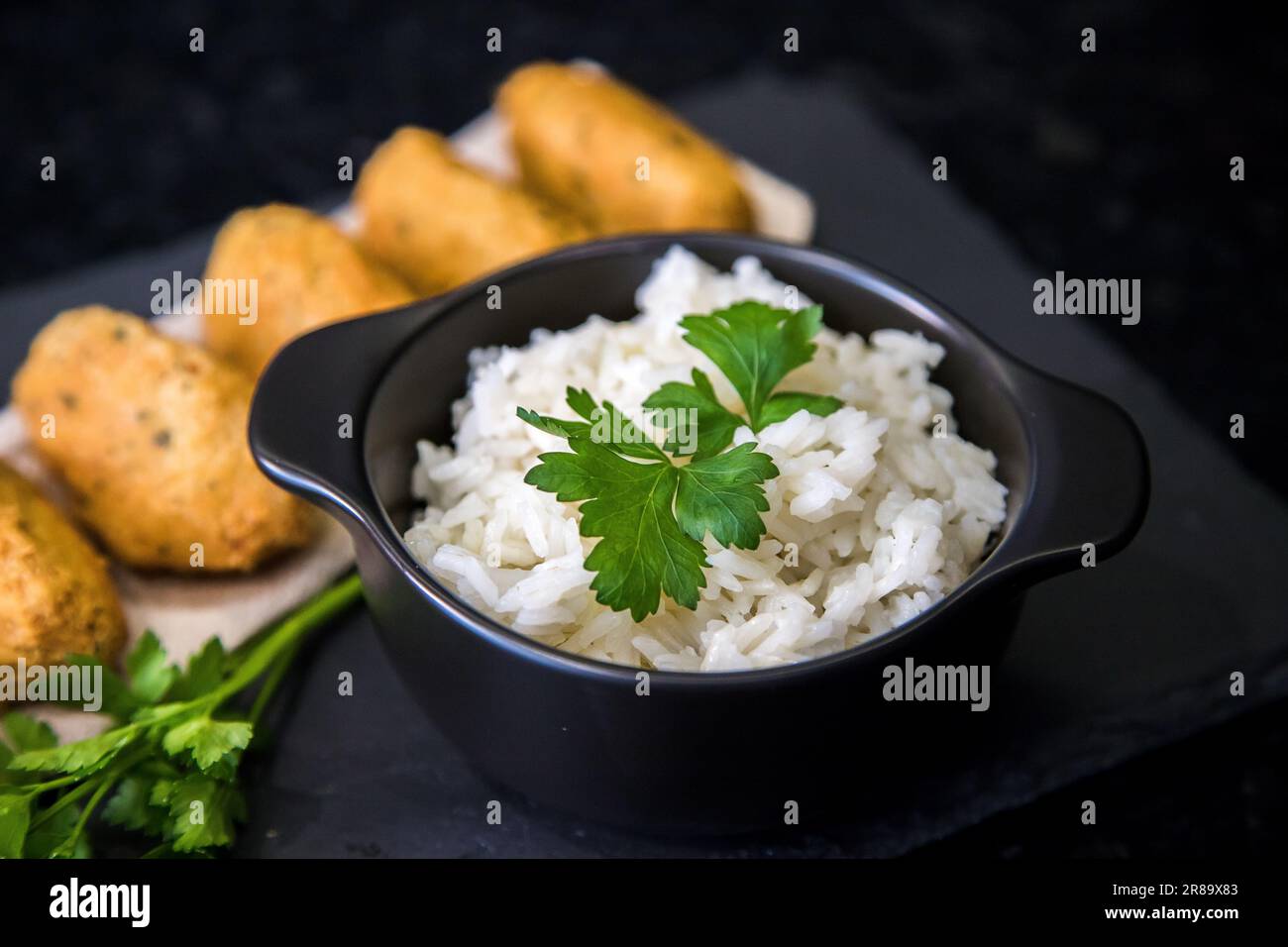 Bolinhos de bacalhau, sehr berühmt in der portugiesischen Gastronomie. Kabeljaukuchen serviert mit weißem Reis auf dunklem Hintergrund. Stockfoto