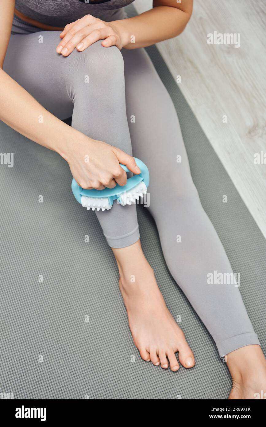 Draufsicht einer jungen Barfußfrau in Aktivbekleidung, Massageunterlage, Bein mit Griff, während sie zu Hause auf einer Fitnessmatte auf dem Boden sitzt, innere Ruhe und Schaden Stockfoto