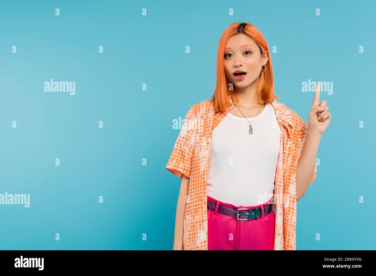 Zeige mit dem Finger, erstaunt, junge asiatische Frau mit gefärbtem Haar, die in die Kamera schaut und etwas auf blauem Hintergrund zeigt, Generation z, zwanglose A Stockfoto