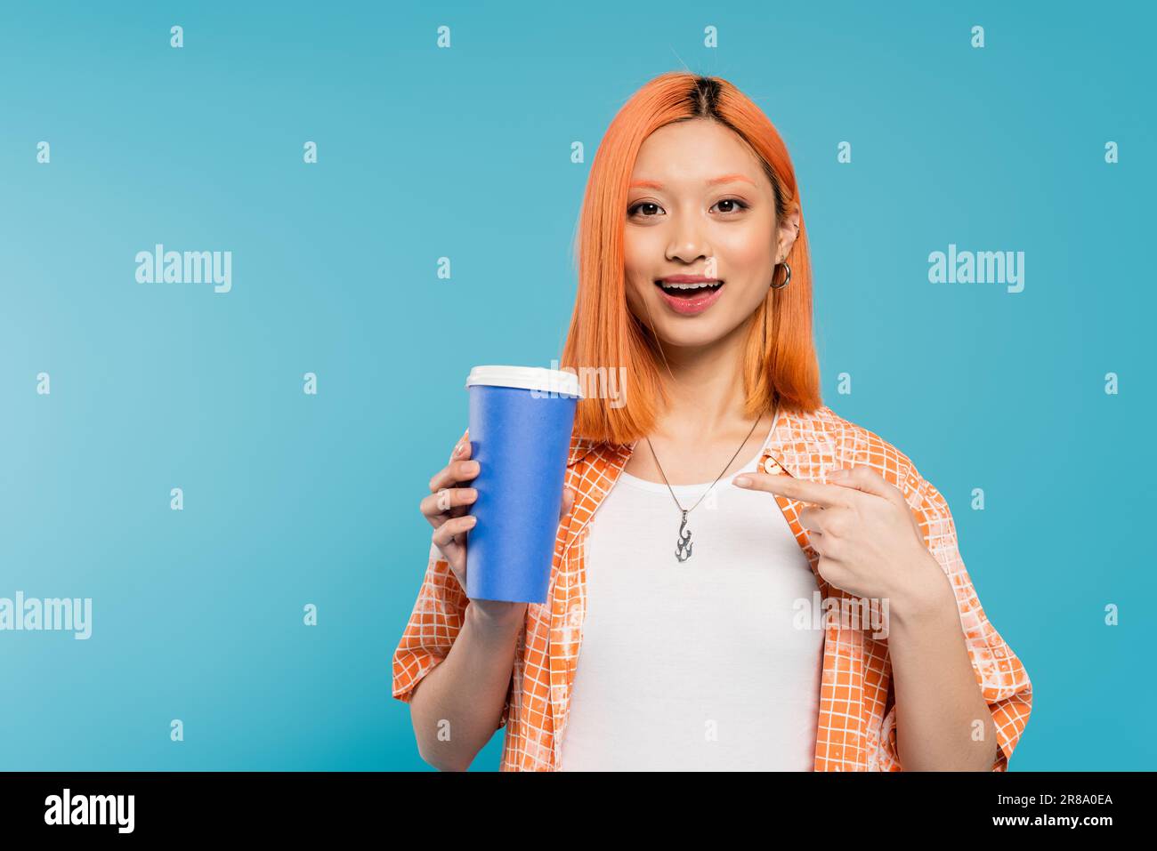 Positive Einstellung, glückliche Asiatin und junge Frau mit roten Haaren, die auf den Pappbecher zeigen und auf die Kamera schauen, auf blauem Hintergrund, Freizeitkleidung, Generation z, Cof Stockfoto