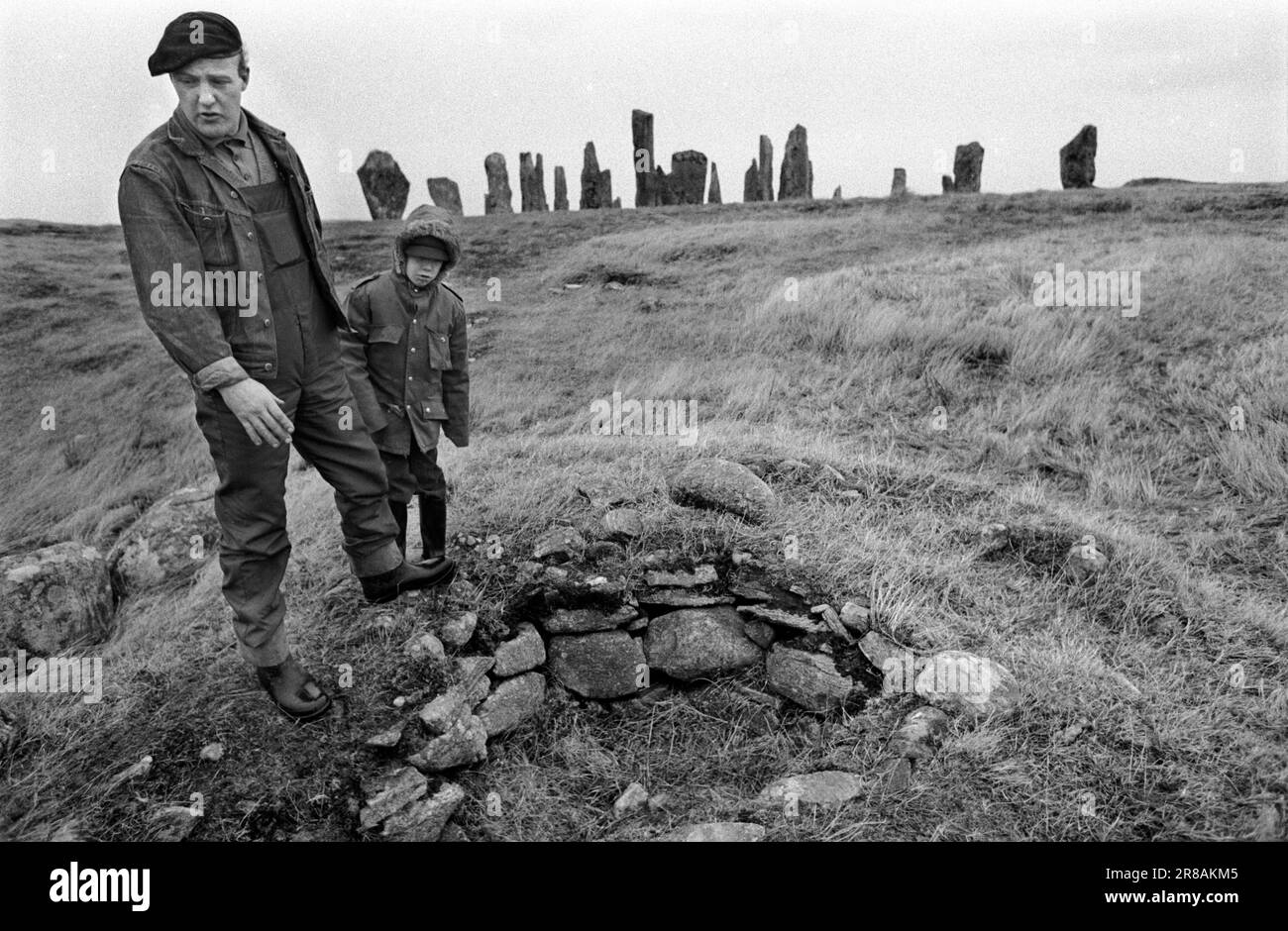 Callanish Standing Stones, Isle of Lewis, Outer Hebrides Schottland, ca. Juni 1974. Die Callanish Stones sind eine Anordnung von Steinen in einem Kreuzmuster mit einem zentralen Steinkreis. Sie wurden in der späten Jungsteinzeit errichtet und waren ein Schwerpunkt für rituelle Aktivitäten während der Bronzezeit. Vater und Sohn der Dorfbewohner weisen auf einen liegengebliebenen Begräbniszist hin - einen kleinen, aus Stein gebauten Sarg. 1970S GB HOMER SYKES Stockfoto