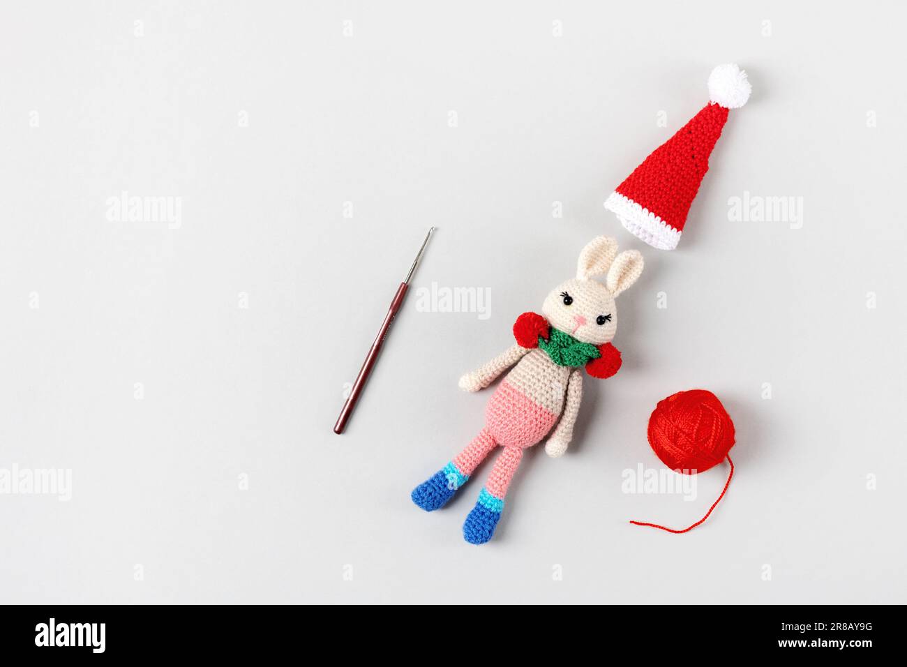 Gestricktes Spielzeughasen mit roter Mütze und Schal mit Fäden und Strickhaken auf grauem Hintergrund. Babysachen und Accessoires. Geschenk für den Winterurlaub Stockfoto