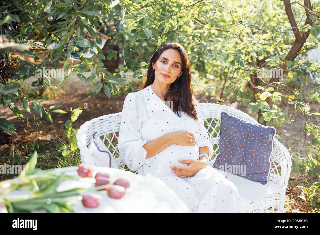 Die charmante, schwangere Brünette in elegantem Kleid sitzt auf einem weißen Rattansofa mit Kissen im Garten. Junge Frau, die ein Baby erwartet, hält sanft h Stockfoto