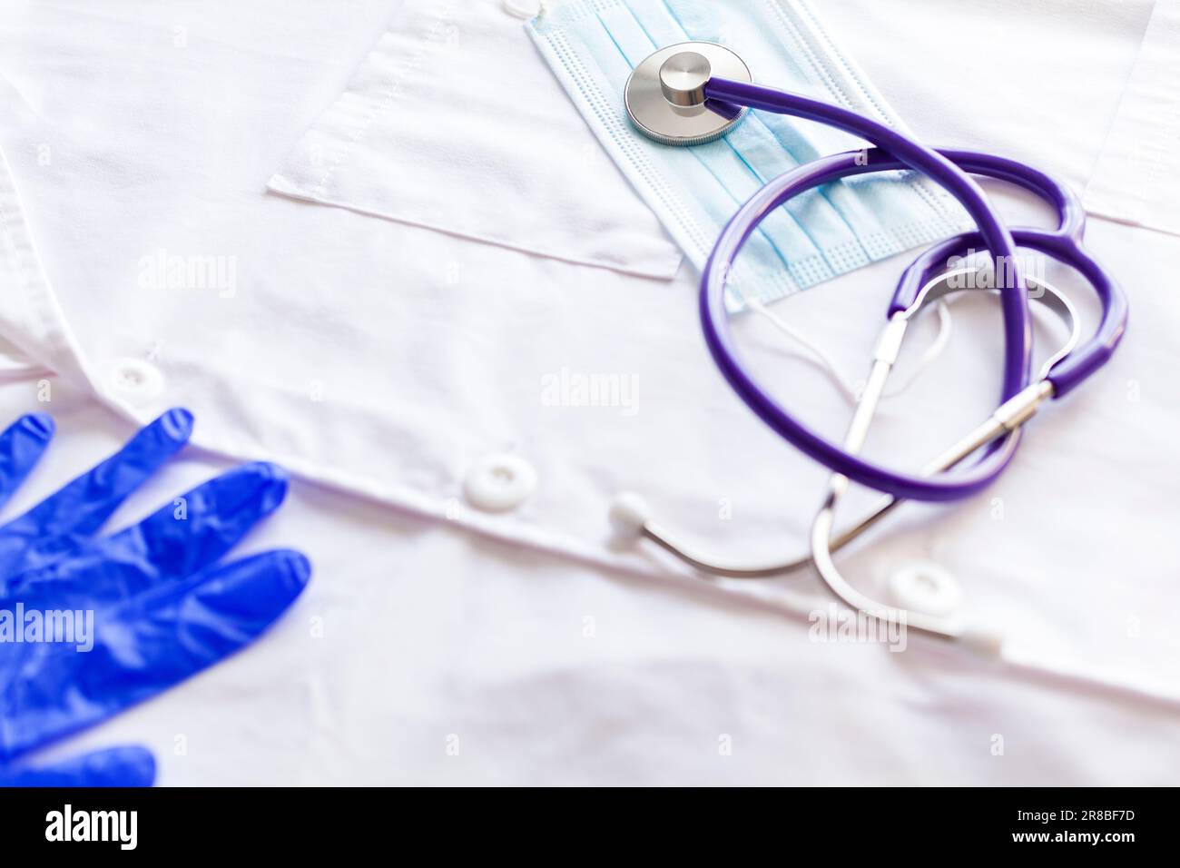 Nahaufnahme eines Stethoskops und einer Operationsmaske über einem weißen medizinischen Kittel und einem Teil eines blauen Latexhandschuhs. Stockfoto