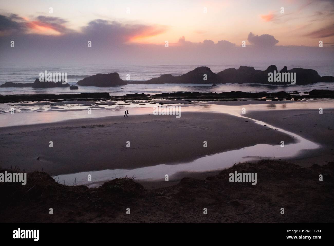 Ein abgelegenes Paar, das an der Küste Oregons entlang spaziert und dabei einen violett-pinkfarbenen Sonnenuntergang mit Seestöcken am Horizont genießt. Stockfoto