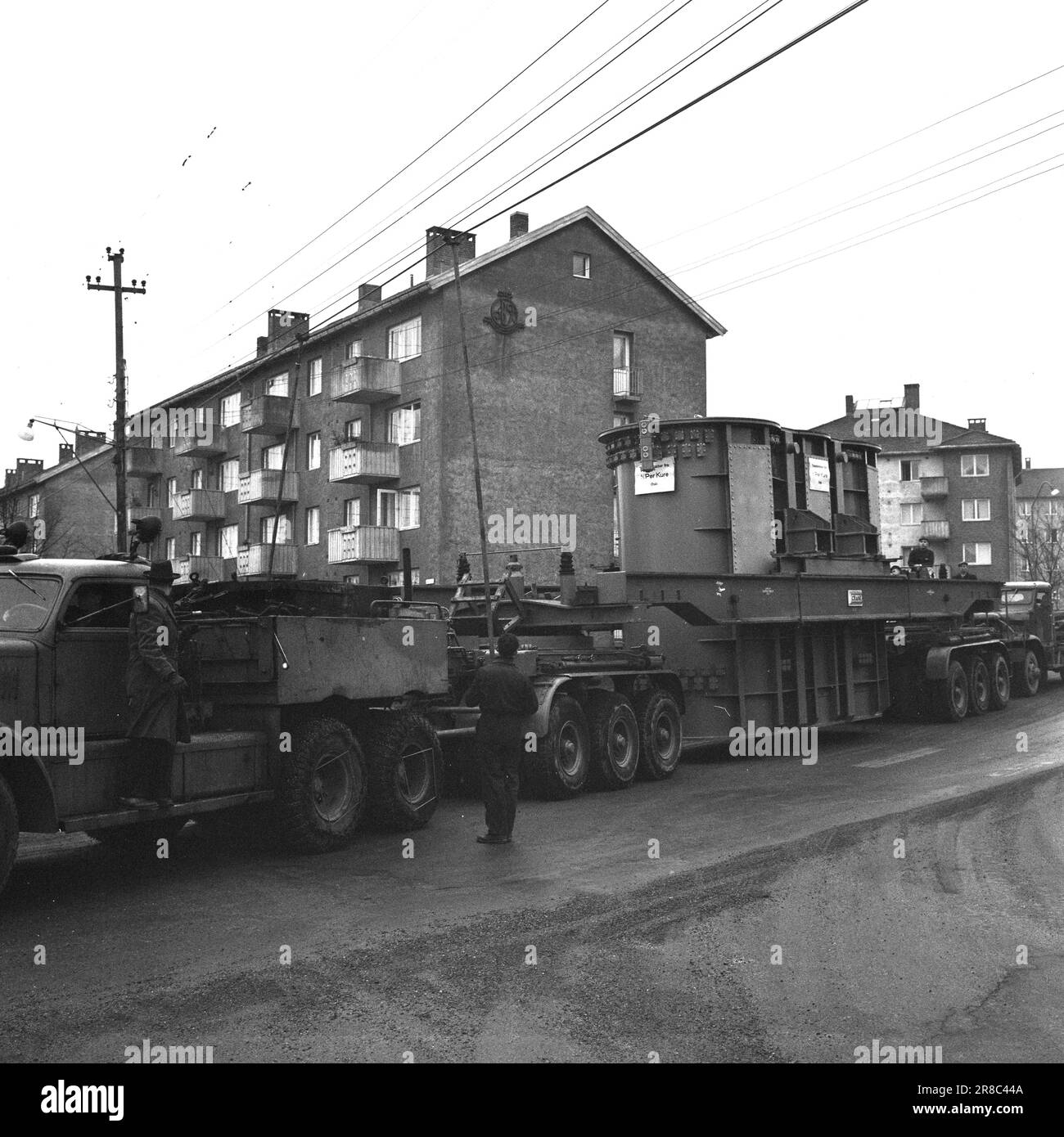 Aktuell 57-3-1960: Riese auf Stadtbesichtigung vierzig Räder, die neulich gemeinsam von Hasle nach Ulven in Oslo gerollt wurden. Sie führten eine historische Aufgabe aus: Der bisher größte in Norwegen gebaute Transformator – ein Riese von 205 Tonnen – wurde von per Kure A/S zum Standort der Beleuchtungsfirma in Ulven transportiert, wo er bei der Stromversorgung helfen wird. Der Transformator, der 161.000 Kilovoltampère erzeugt, ist 9,6 Meter lang und 5,7 Meter breit. Oslo Lysverker hatte Verstärkung für den Großtransport angefordert, und nicht weniger als zwei Fahrer waren im Geschäft. Sie erhielten ihre Fahrbefehle vom Komma Stockfoto