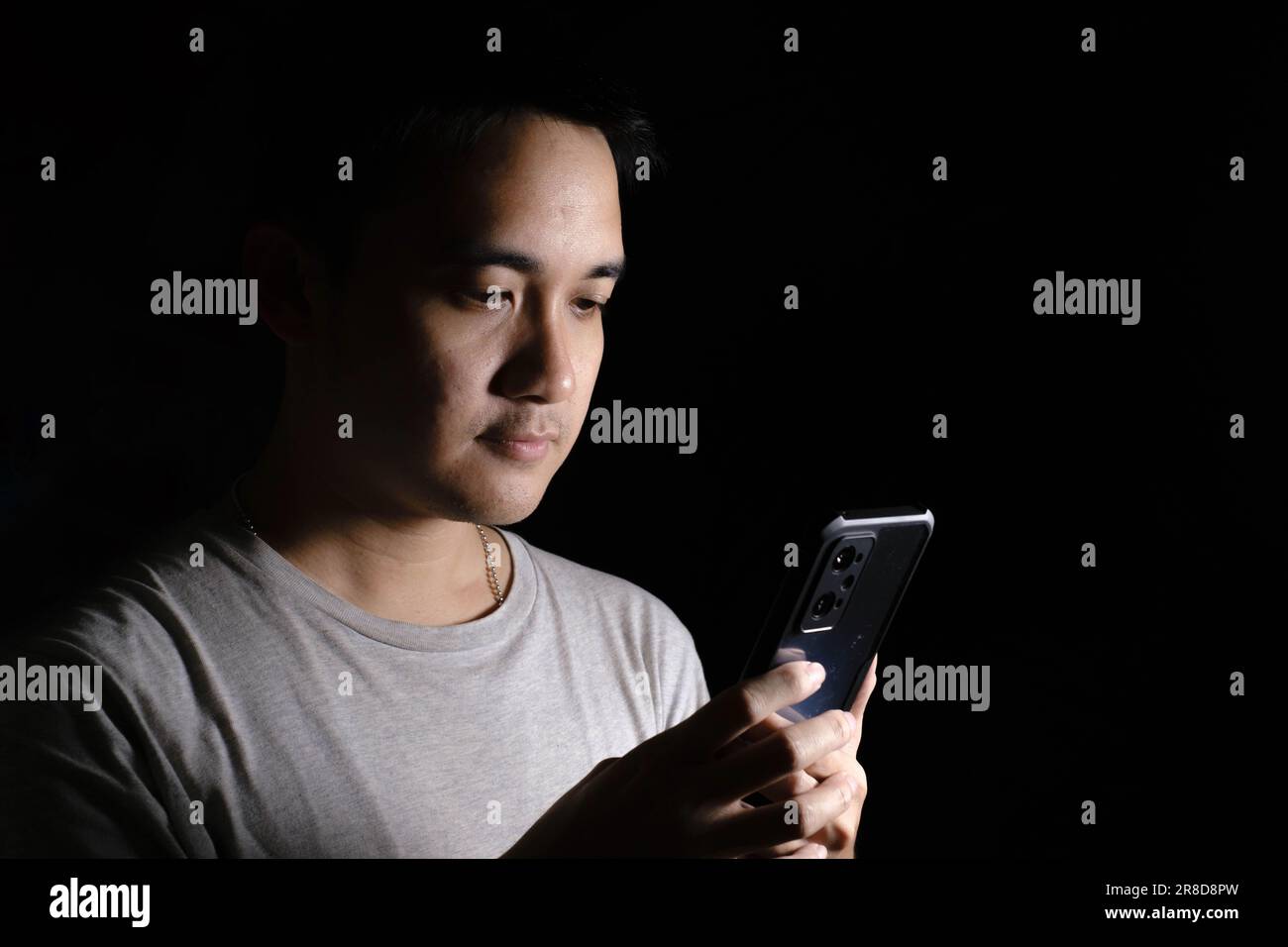 Unauffällige Aufnahme eines jungen Asiaten, der ein graues T-Shirt trägt und auf ein Smartphone schaut. Isolierter schwarzer Hintergrund. Dramatisch. Stockfoto