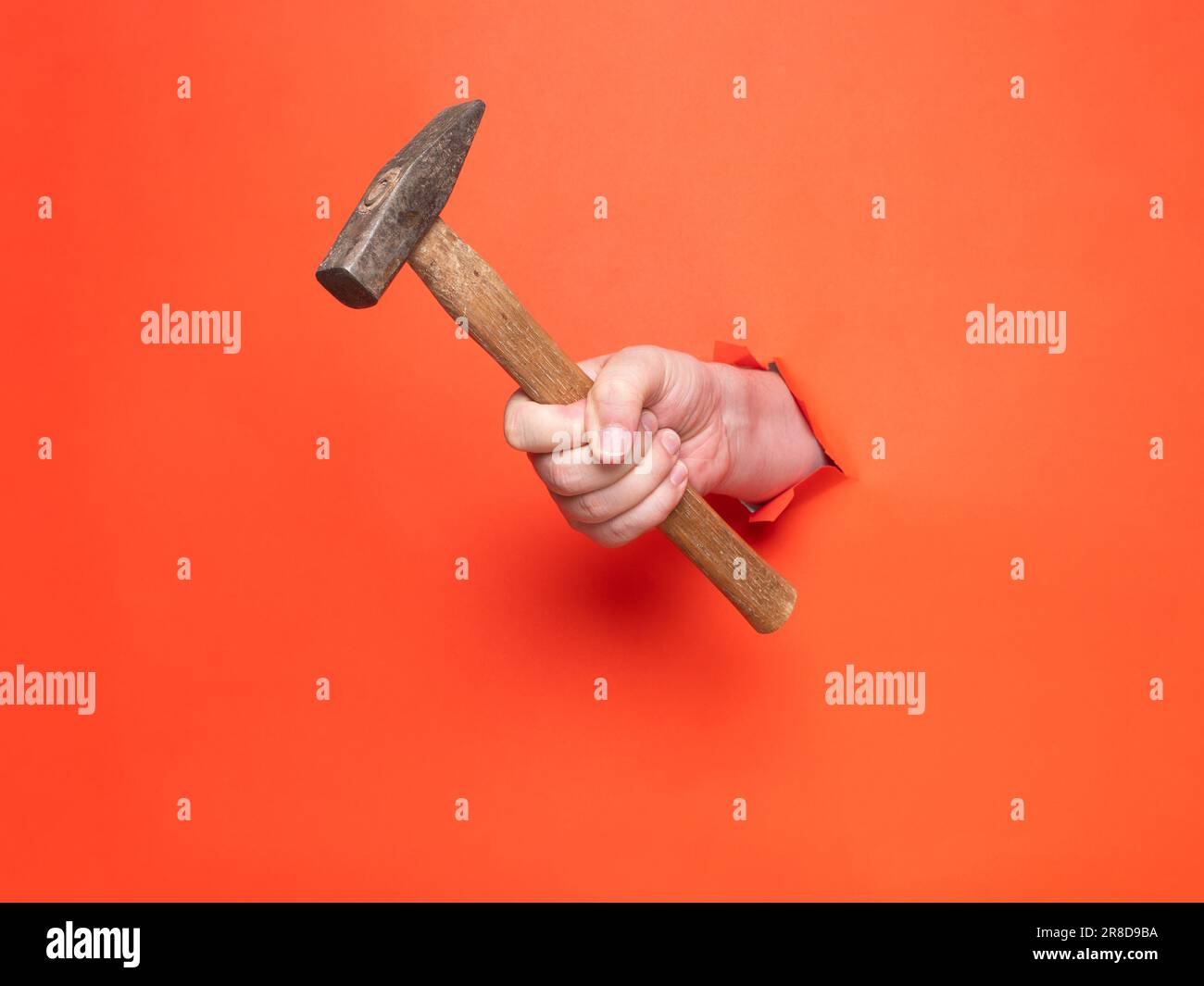 Die Hand eines Mannes hält einen Hammer durch zerrissenes orangefarbenes Papier. Konzept, Hand durch zerrissenes orangefarbenes Papier. Stockfoto
