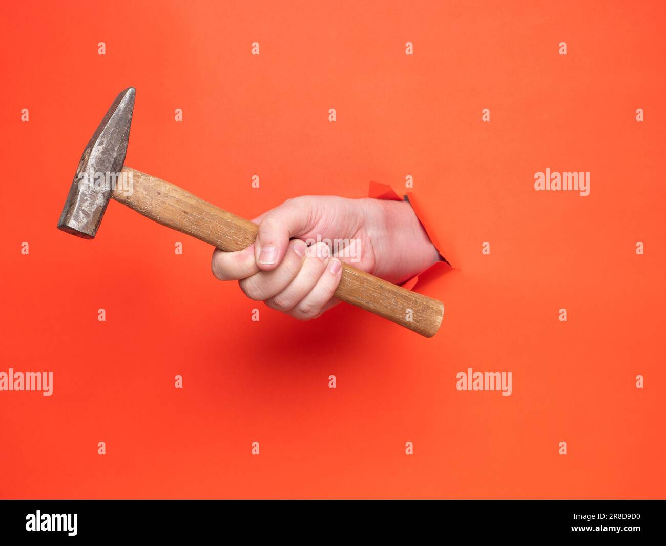 Die Hand eines Mannes hält einen Hammer durch zerrissenes orangefarbenes Papier. Konzept, Hand durch zerrissenes orangefarbenes Papier. Stockfoto