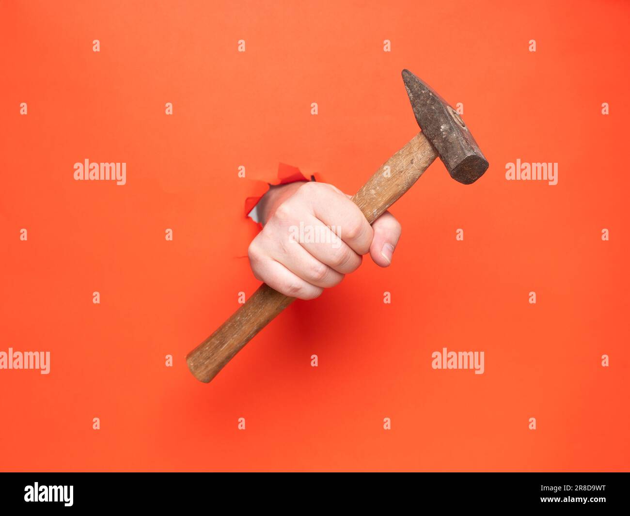 Die Hand eines Mannes hält einen Hammer durch zerrissenes orangefarbenes Papier. Konzept, Hand durch zerrissenes orangefarbenes Papier. Stockfoto