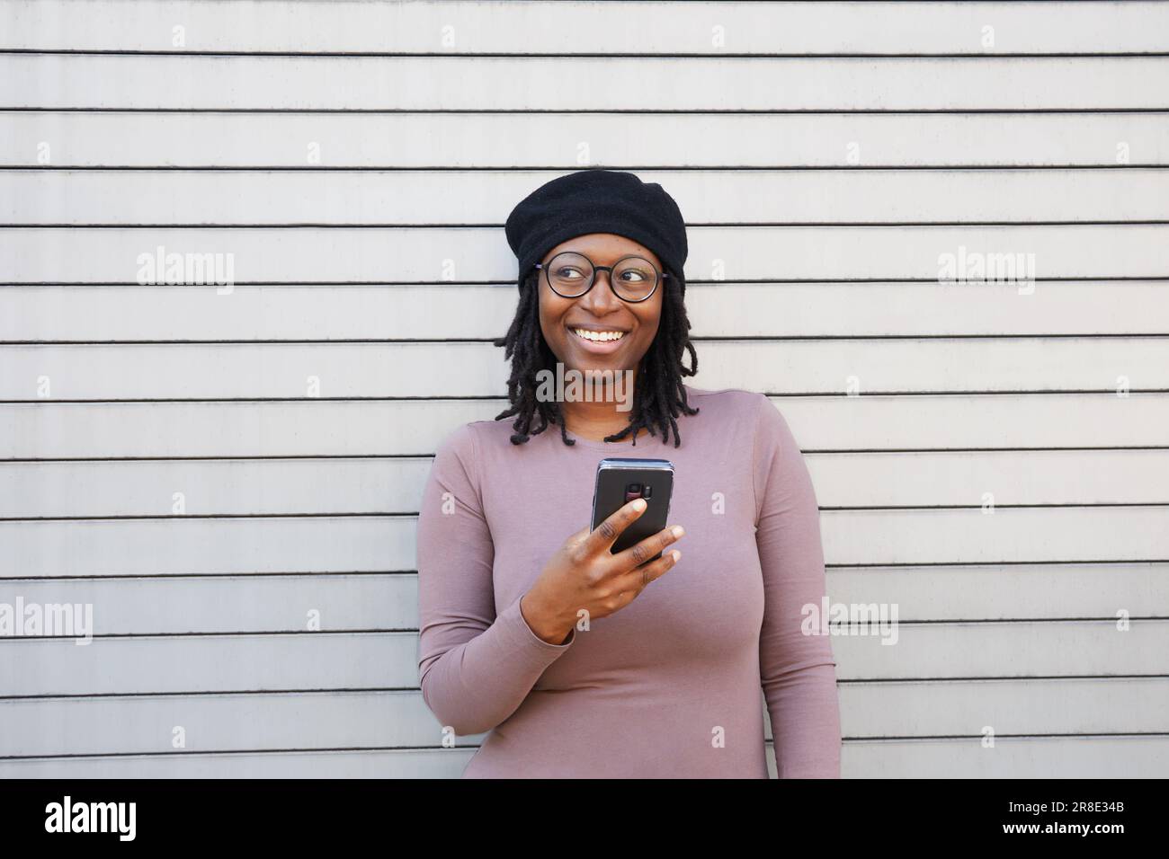 Lächelnde Frau mit Brille und Baskenmütze mit Smartphone in der Stadt Stockfoto