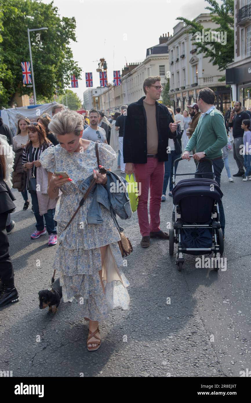 Frau, die einen Hund begleitet und gleichzeitig ein iphone liest. Die Kings Road, Platinum Jubilee Street Party, die den Aufruf zur Krise des britischen Roten Kreuzes in der Ukraine unterstützt. Die Kings Road ist zum Teil für den Verkehr gesperrt, lokale Chelsea und Knightsbridge junge Erwachsene unterhalten sich, während eine Frau mit ihrem Hund geht und liest oder iphone. Chelsea, London, England 4. Juni 2022. HOMER SYKES AUS DEN 2020ER JAHREN Stockfoto