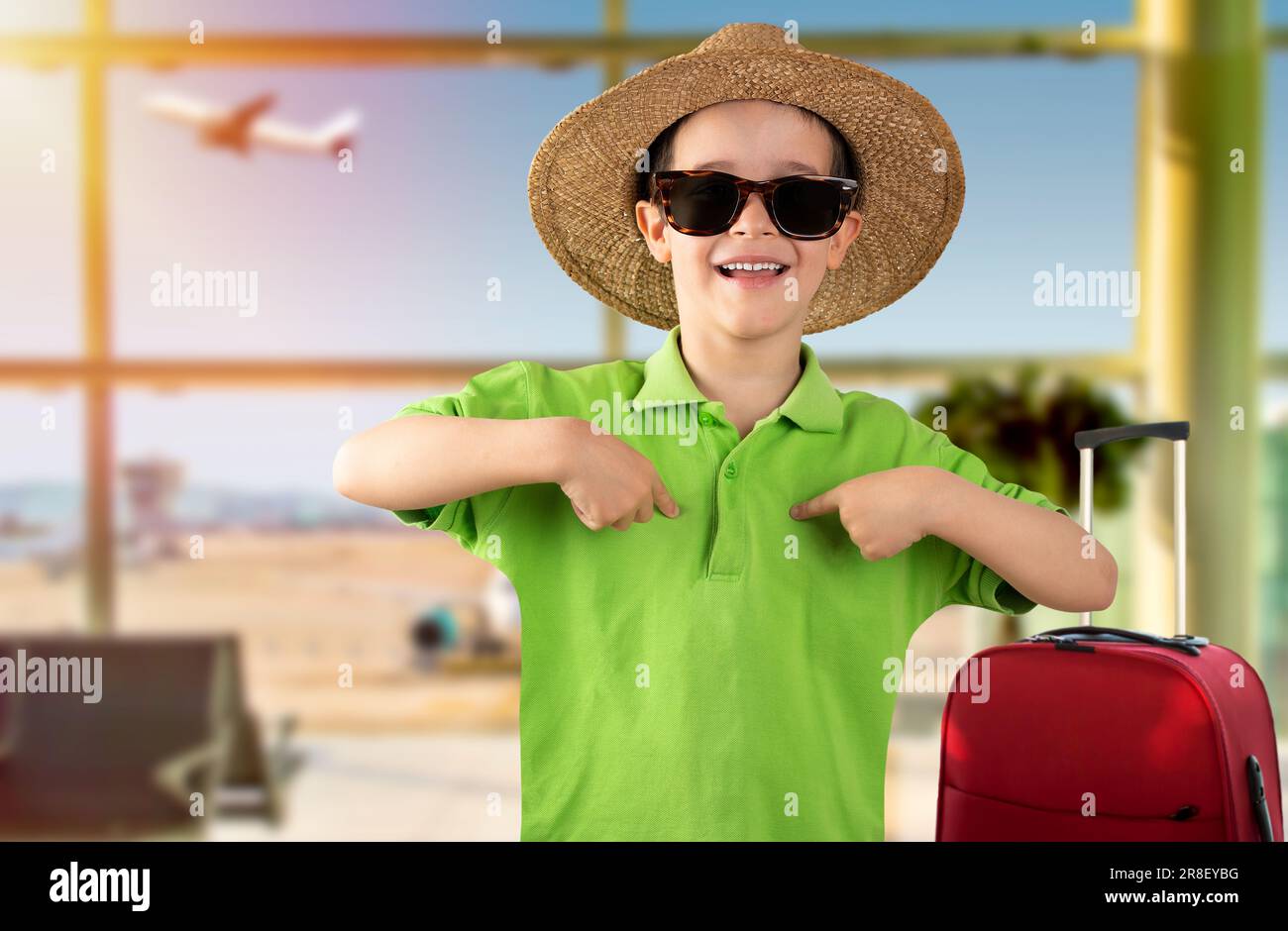 Ein Kind im Urlaub trägt eine Sonnenbrille mit grünem T-Shirt und Hut am Flughafen und sieht selbstbewusst mit einem Lächeln im Gesicht aus, zeigt sich mit stolzen Fingern und ha Stockfoto