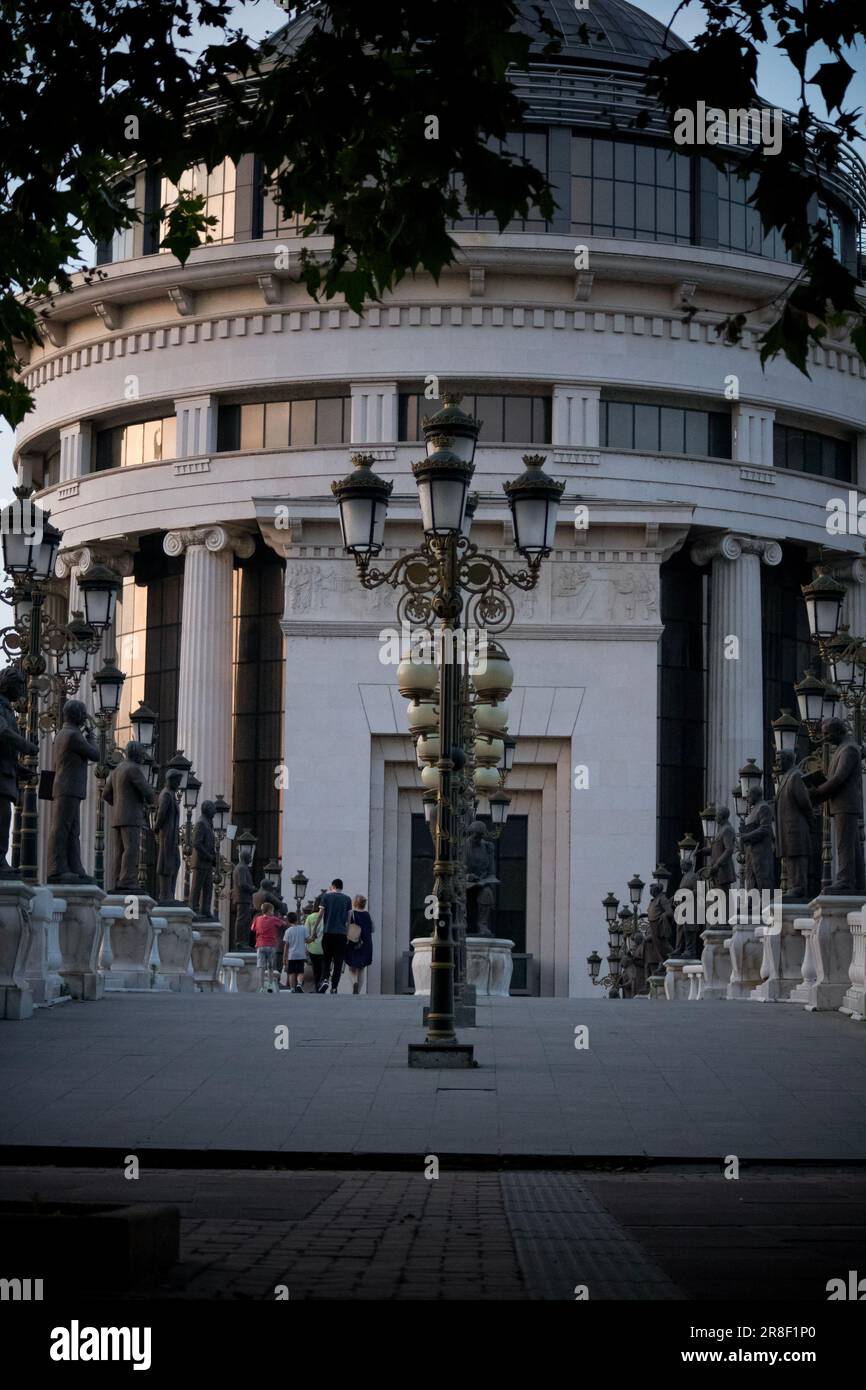 Kunstbrücke mit Kupferstatuen und Straßenlaternen auf dem mazedonischen Skopje Stadtplatz, die zu einem Gebäude mit Glas und Reflexion führt, alles während des Sonnenuntergangs Stockfoto