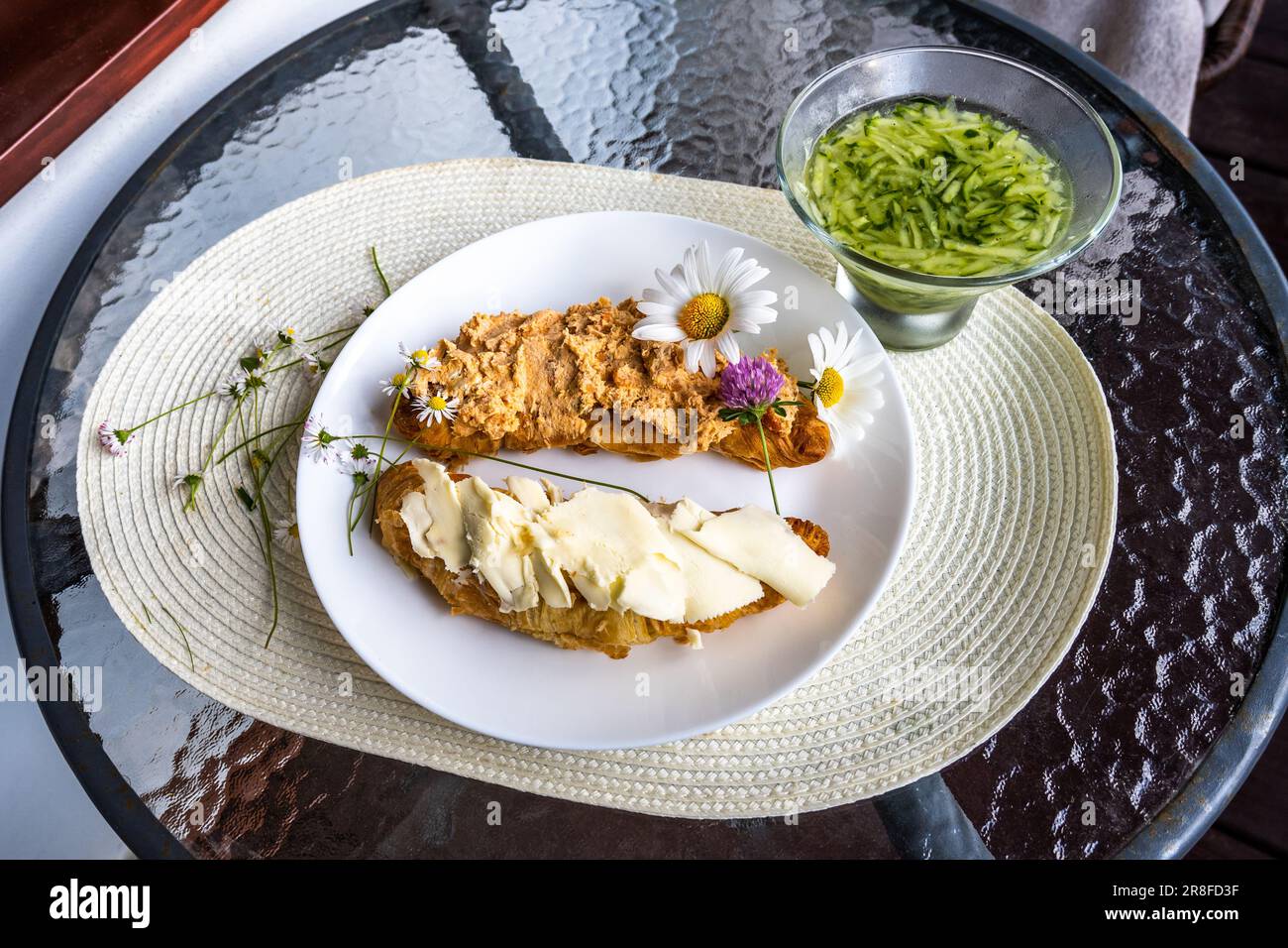 Zwei Stück Croissant mit Butter und Thunfisch auf weißem Teller, Blumen auf Teller gestreut, Gurkensalat auf rundem Glastisch. Gesundes Frühstück o Stockfoto