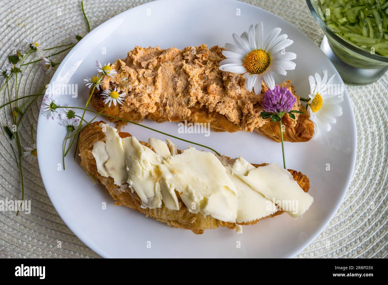 Zwei Stück Croissant mit Butter und Thunfisch auf weißem Teller, Blumen, ein Teil Gurkensalat auf dem Tisch. Gesundes Frühstück oder Snack, Nahaufnahme. Stockfoto