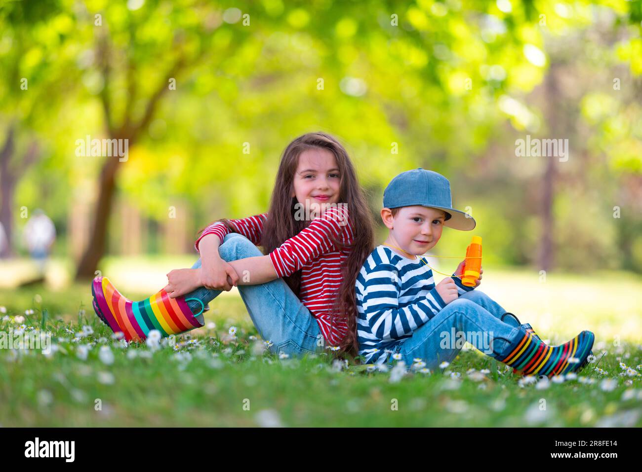 Fröhliche Kinder Junge und Mädchen in Gummistiefeln spielen draußen im grünen Park mit blühenden Blüten Stockfoto