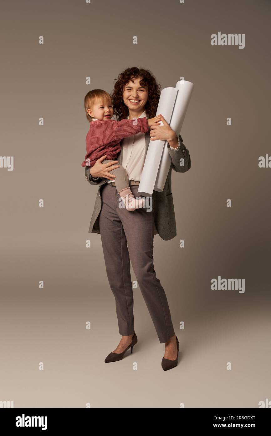 Multitasking-Frau, berufliche Leistungen, Zeitmanagement, glückliche Mutter, die sich in den Armen hält Tochter und gerollte Papierpläne auf grauem Hintergrund, Stockfoto