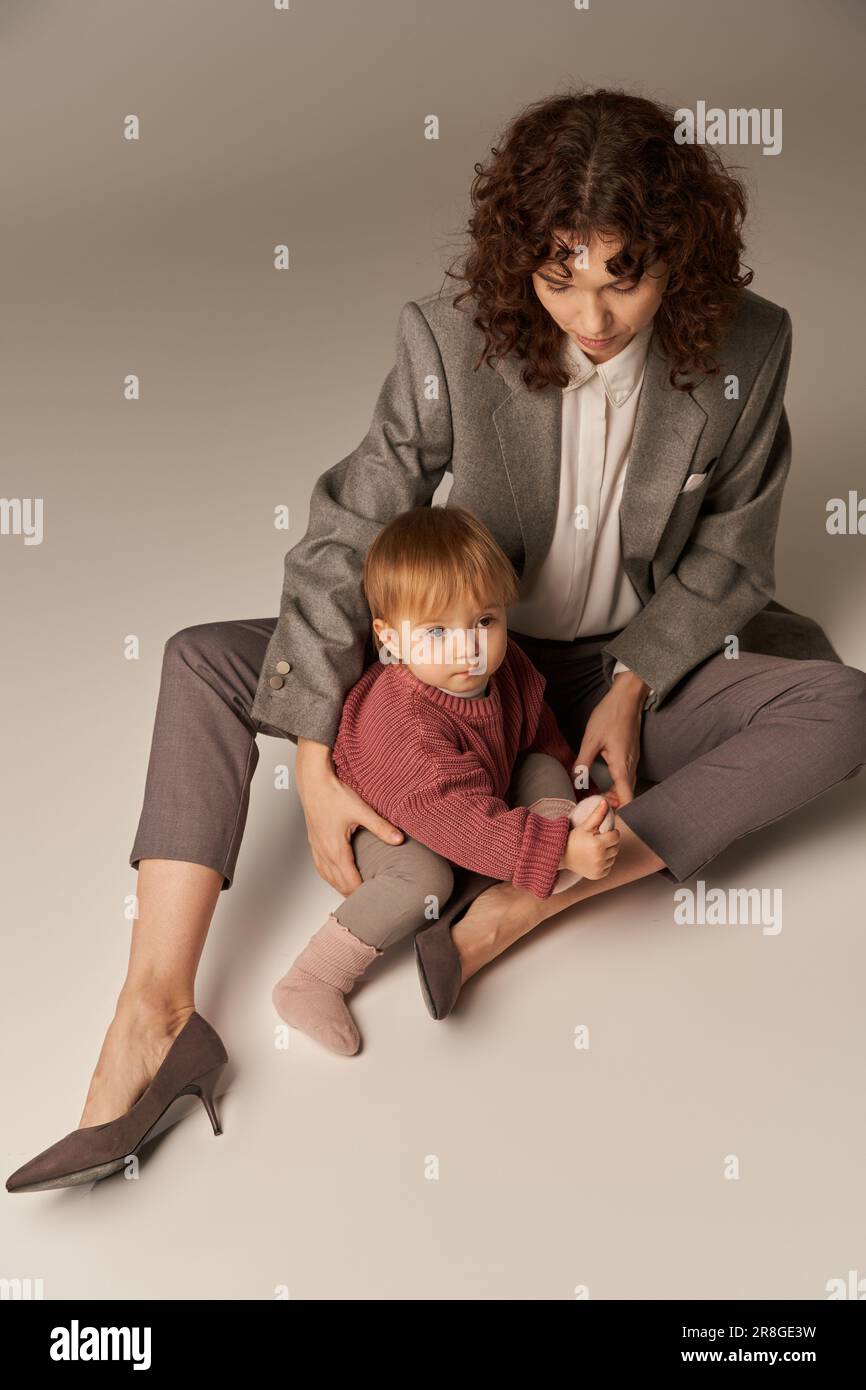 Karriere und Familie, Gleichgewicht zwischen Arbeit und Leben, fröhliche Mutter mit lockigem Haar umarmte Kleinkinder Tochter auf grauem Hintergrund, schöne Zeit, modern Stockfoto