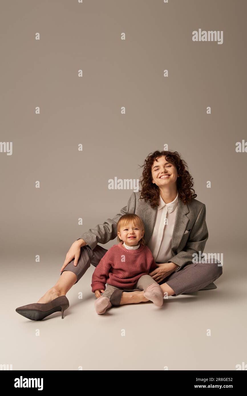 Karriere und Familie, Gleichgewicht zwischen Arbeit und Leben, glückliche Frau mit lockigem Haar, die kleine Tochter auf grauem Hintergrund umarmt, schöne Zeit, modernes Pare Stockfoto