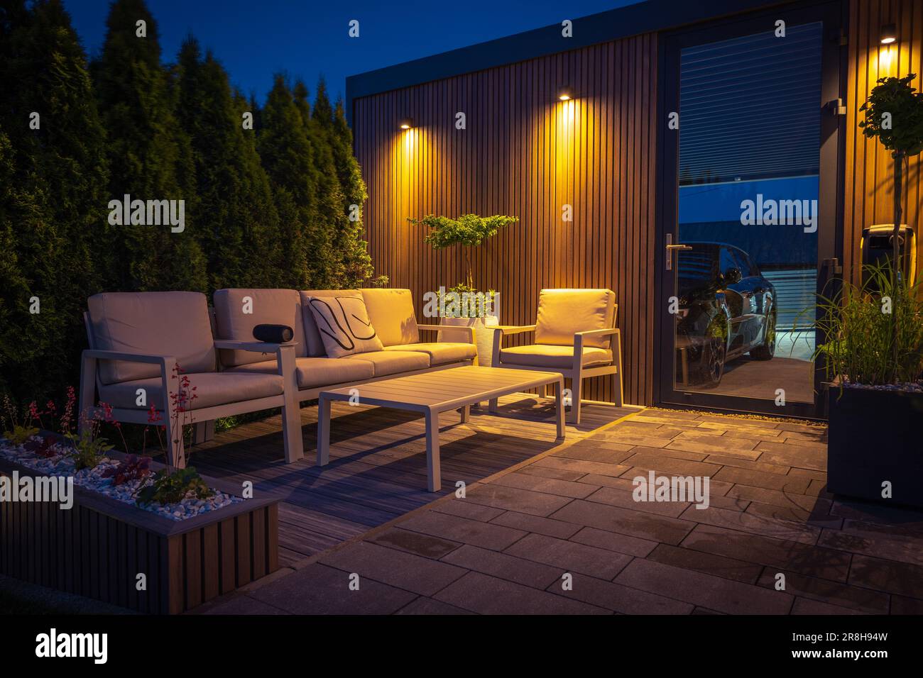 Lounge- und Essbereich im modernen Hinterhof mit Außenbeleuchtung, Pflanzen, Gartentisch und Stühlen. Gemütlicher Sommerabend. Stockfoto