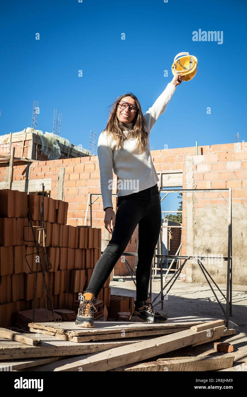 Erfolgreiche, reife Ingenieurin. Vertikales Porträt einer Architektin, die lächelt und in die Kamera schaut, mit ihrem gelben Schutzhelm, der sie hochhält Stockfoto