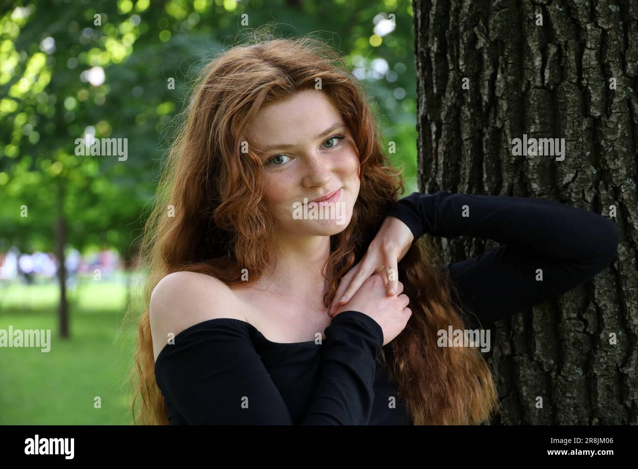 Attraktives lächelndes Mädchen mit langen roten Haaren und Sommersprossen, das neben dem Baum im Sommerpark steht. Junges Model mit schwarzem Oberteil, weibliche Schönheit Stockfoto