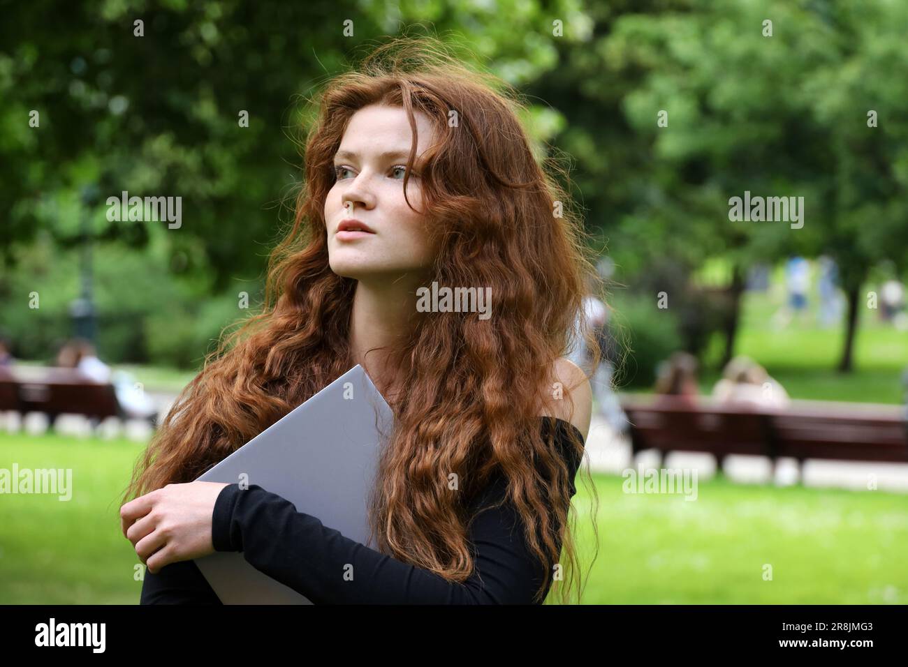 Inspirierendes Mädchen mit langen roten Haaren und Sommersprossen, das im Sommerpark mit einem Laptop in der Hand steht. Lernen und Kreativität in der Natur Stockfoto