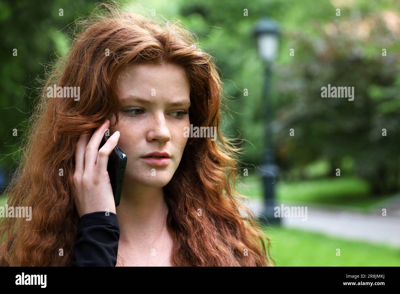 Attraktives Mädchen mit langen roten Haaren und Sommersprossen, das im Sommerpark telefoniert Stockfoto