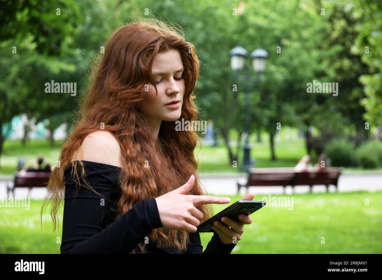 Lächelndes Mädchen mit langen roten Haaren und Sommersprossen, das im Sommerpark auf dem Handy steht. Konzept von Flirten und Online-Dating Stockfoto