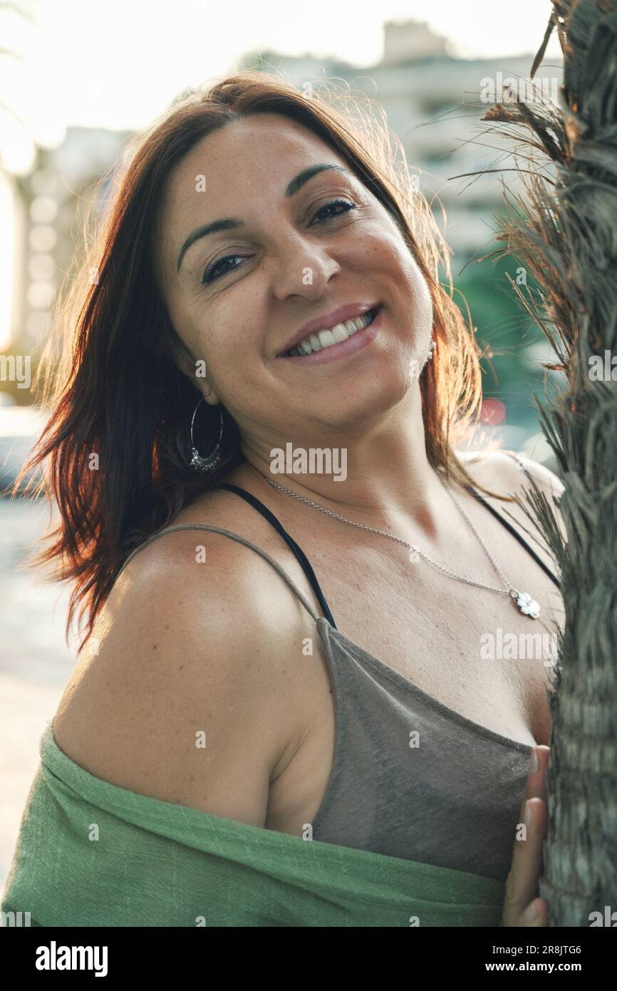 Lebendiges Porträt einer lächelnden brünetten Frau, die sich gegen eine Palme lehnt und Authentizität und natürliche Schönheit in Reise- und Lifestyle-Bildern festhält. Stockfoto