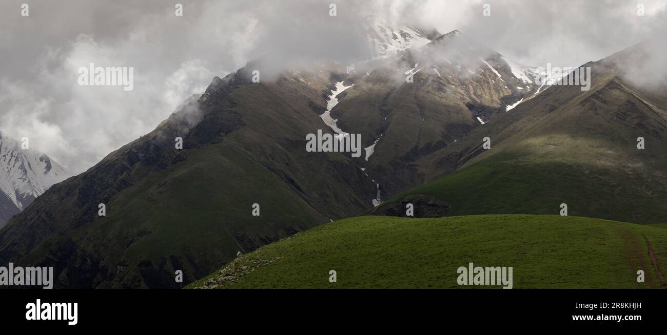 Bergpanorama. Berggipfel in den Wolken, eine Bergwiese, ein Wasserfall in einer Bergschlucht. Stockfoto
