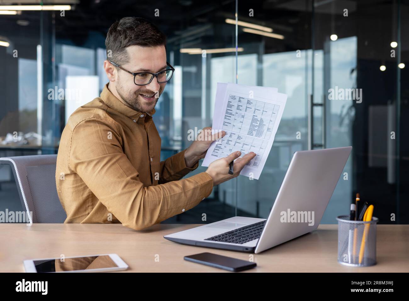 Ein junger lächelnder Geschäftsmann hält ein Online-Geschäftstreffen ab. Er sitzt im Büro am Tisch vor dem Laptop und kommuniziert per Videoanruf, zeigt Dokumente und Berichte auf dem Bildschirm an. Stockfoto