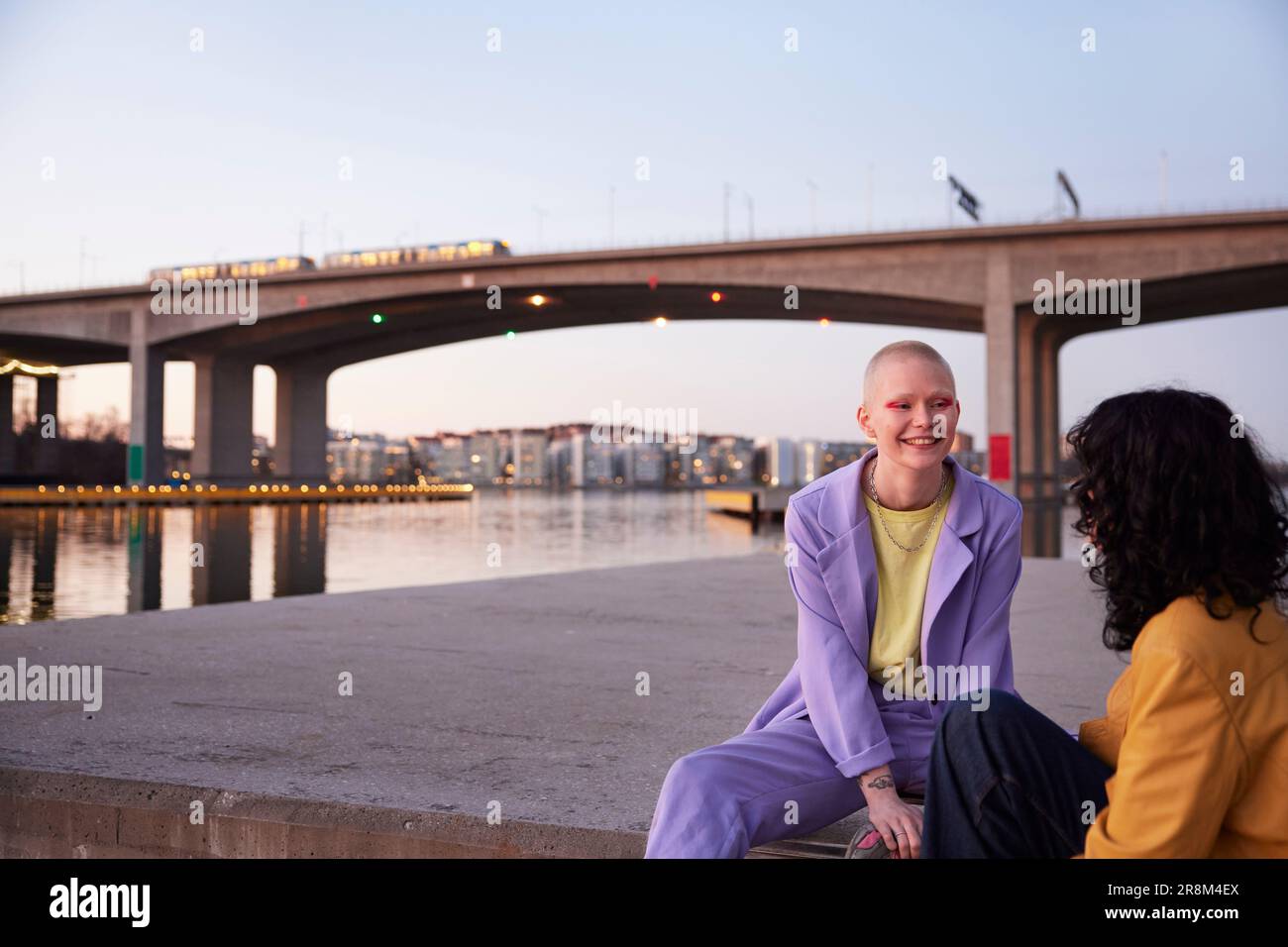 Ein junges weibliches schwules Paar, das am Fluss sitzt und redet, eine Brücke im Hintergrund Stockfoto