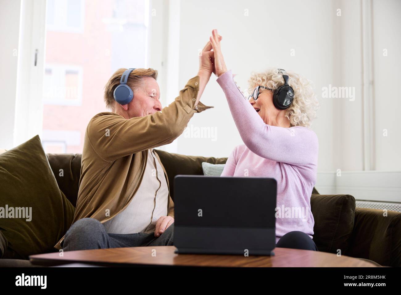 Ältere Männer und Frauen geben sich gegenseitig High Five, während sie im Wohnzimmer vor dem digitalen Tablet sitzen Stockfoto