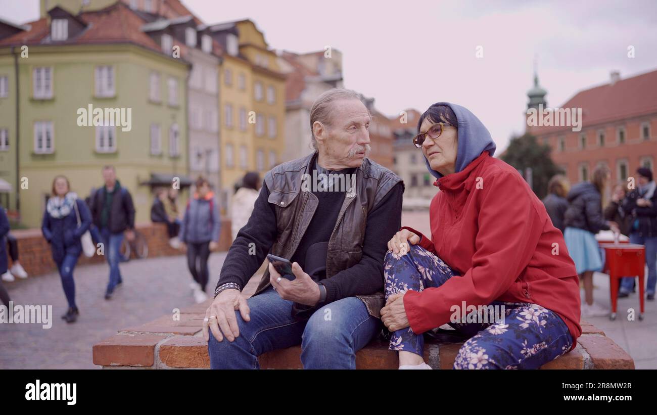 Ältere Touristen sitzen und unterhalten sich im historischen Zentrum einer alten europäischen Stadt. Senior hat ein Smartphone in der Hand. Palast Stockfoto