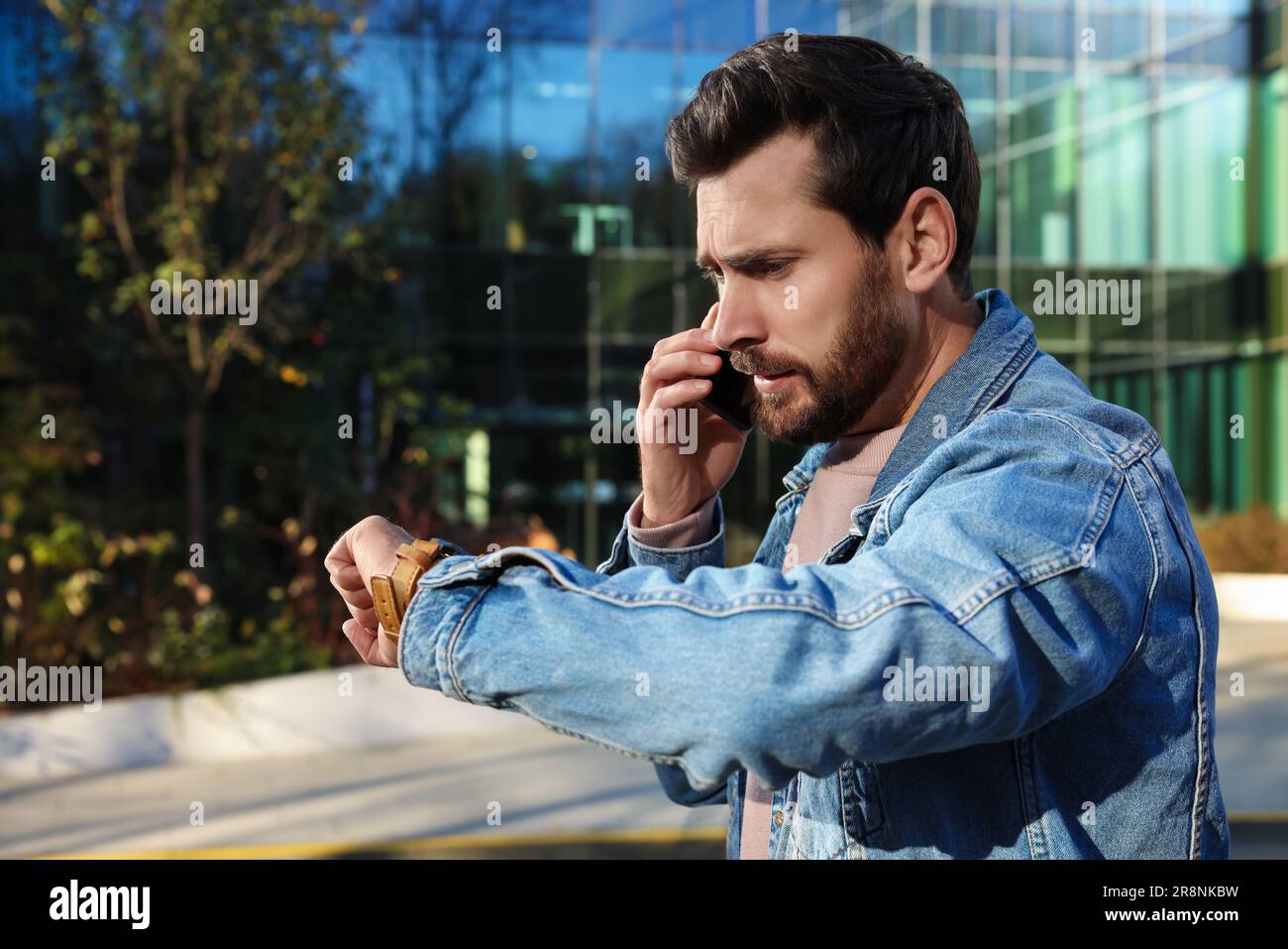 Emotionaler Mann, der auf einem Smartphone spricht und auf eine Uhr in der Nähe eines Gebäudes schaut. Das Konzept der Verspätung Stockfoto