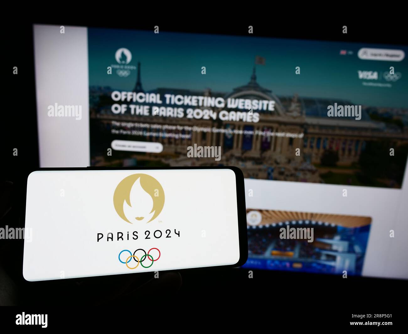 Person, die ein Handy mit dem Logo der Olympischen Sommerspiele 2024 in Paris auf dem Bildschirm vor der Webseite hält. Konzentrieren Sie sich auf das Display des Telefons. Stockfoto