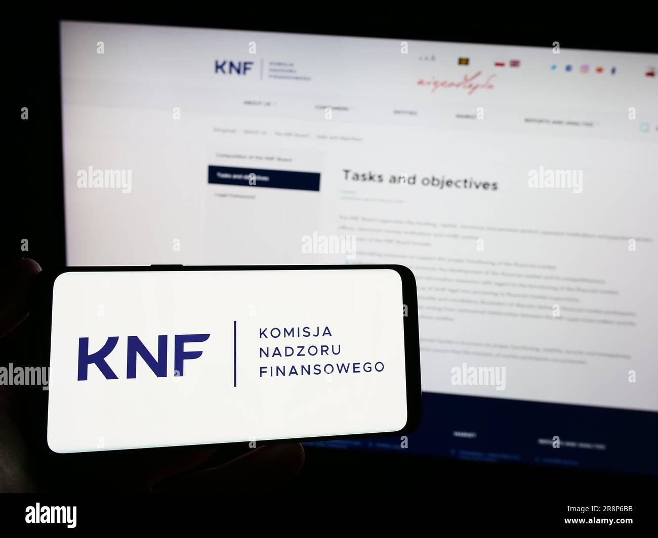 Person, die ein Smartphone mit dem Logo der Behörde Komisja Nadzoru Finansowego (KNF) auf dem Bildschirm vor der Website hält. Konzentrieren Sie sich auf das Display des Telefons. Stockfoto