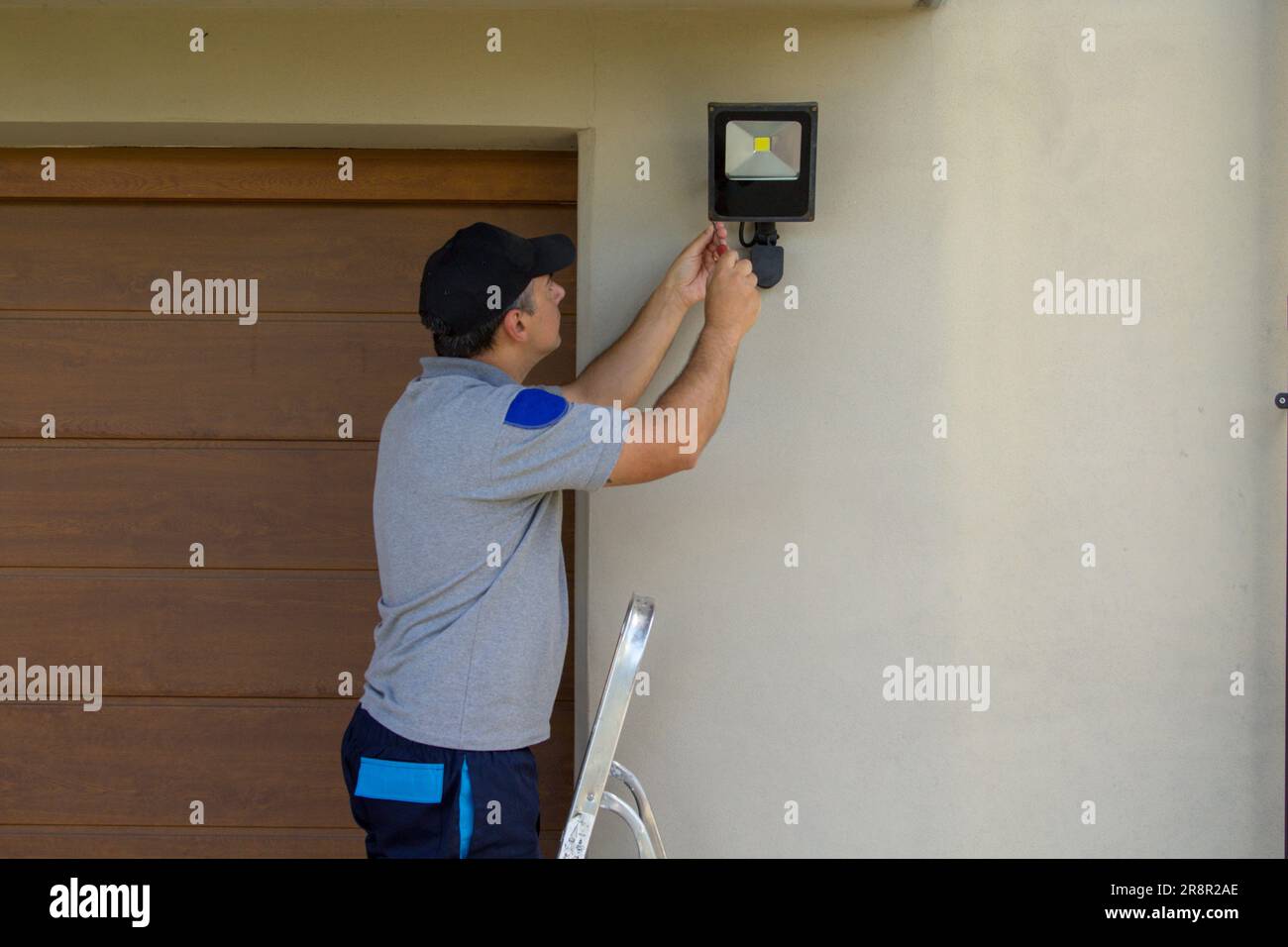 Bild eines Elektrikers auf einer Scherenleiter, der mit seinen Werkzeugen einen Scheinwerfer an einem Haus installiert. Beleuchtung der dunkelsten Teile des Hauses. Stockfoto