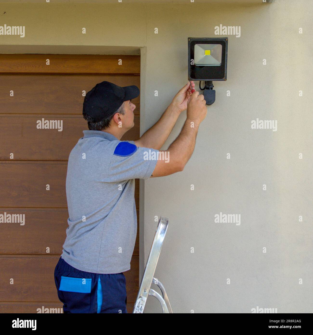Bild eines Elektrikers auf einer Scherenleiter, der mit seinen Werkzeugen einen Scheinwerfer an einem Haus installiert. Beleuchtung der dunkelsten Teile des Hauses. Stockfoto