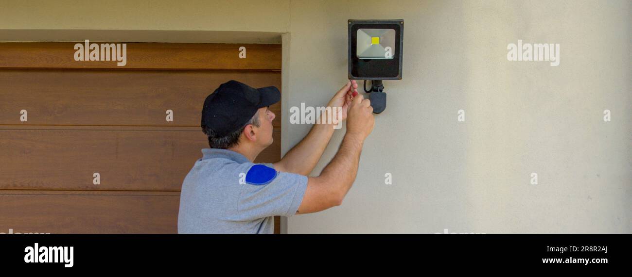 Bild eines Elektrikers auf einer Scherenleiter, der mit seinen Werkzeugen einen Scheinwerfer an einem Haus installiert. Beleuchtung der dunkelsten Teile des Hauses. Banner Stockfoto