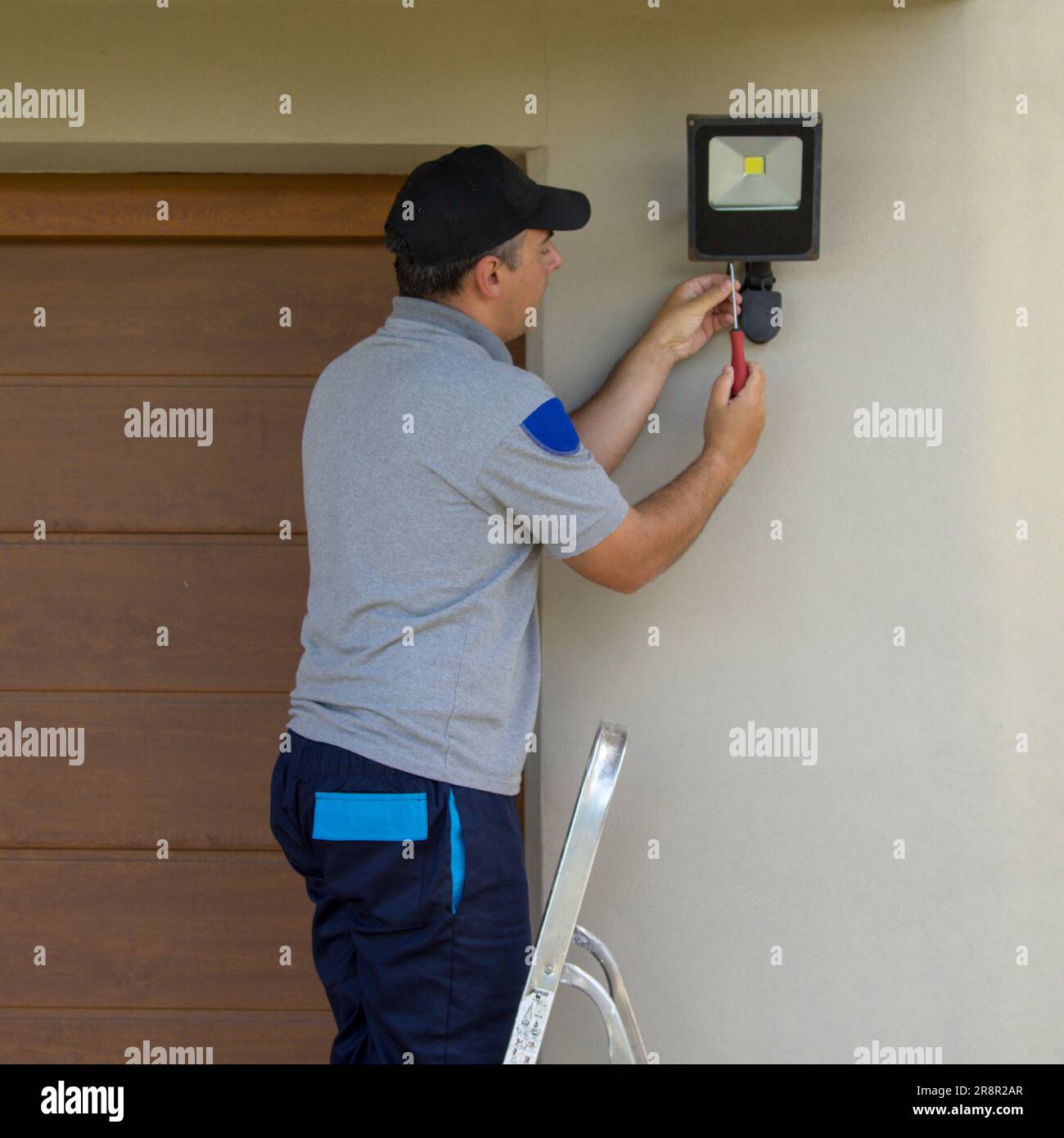 Bild eines Elektrikers auf einer Scherenleiter, der mit seinen Werkzeugen einen Scheinwerfer an einem Haus installiert. Beleuchtung der dunkelsten Teile des Hauses. Stockfoto