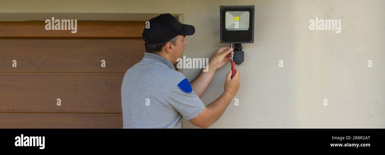 Bild eines Elektrikers auf einer Scherenleiter, der mit seinen Werkzeugen einen Scheinwerfer an einem Haus installiert. Beleuchtung der dunkelsten Teile des Hauses. Banner Stockfoto