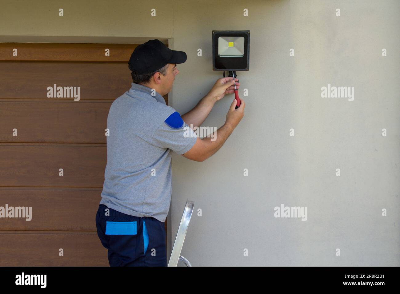 Bild eines Elektrikers auf einer Scherenleiter, der mit seinen Werkzeugen einen Scheinwerfer an einem Haus installiert. Beleuchtung der dunkelsten Teile des Hauses. Stockfoto