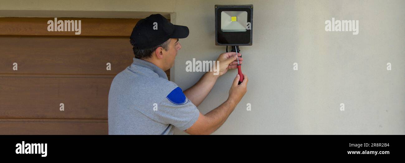 Bild eines Elektrikers auf einer Scherenleiter, der mit seinen Werkzeugen einen Scheinwerfer an einem Haus installiert. Beleuchtung der dunkelsten Teile des Hauses. Banner Stockfoto