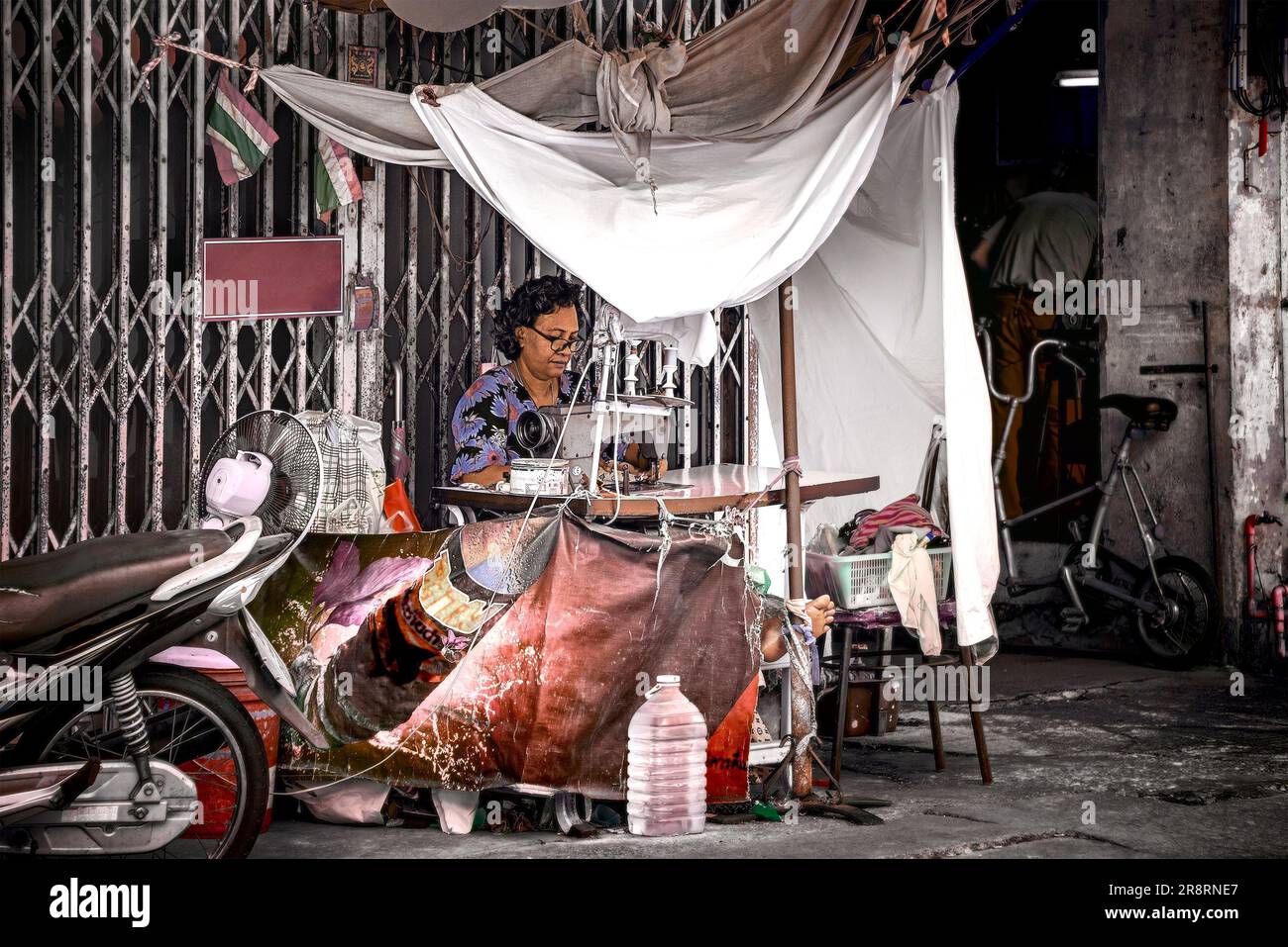 Bangkok, Thailand, 20. März 2023: Straßenkleiderin arbeitet vor einer Falttür aus Stahl und unter einem abgenutzten Sonnenschirm aus Stoff. Stockfoto